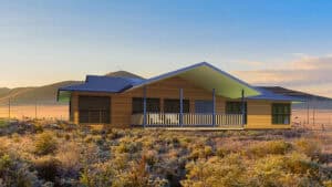 Elevated bungalow with front porch in dry grassland, hills and clear sky in the background.