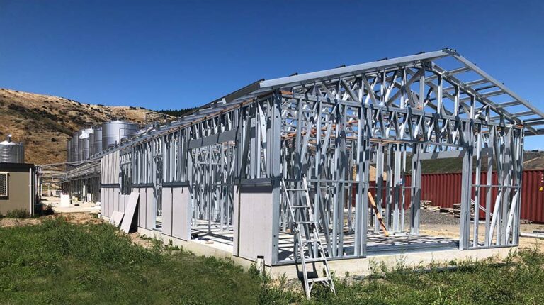 Steel frame construction of a modern greenhouse under development, featuring durable metal structure and wooden accents, set in a scenic rural area with clear skies and rolling hills.