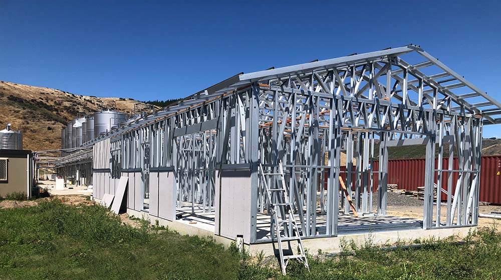 Steel frame construction of a modern greenhouse under development, featuring durable metal structure and wooden accents, set in a scenic rural area with clear skies and rolling hills.