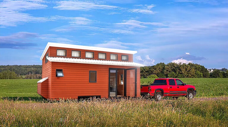 Compact red tiny house on wheels with modern design, large windows, and a small porch, parked in a rural field under a blue sky.