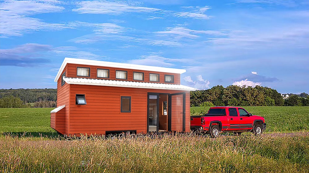 Compact red tiny house on wheels with modern design, large windows, and a small porch, parked in a rural field under a blue sky.