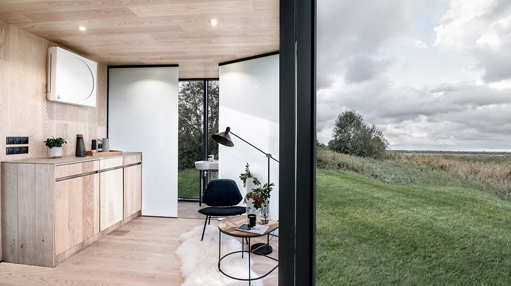 Interior view of a modern mirror cabin with wooden accents, a black chair, and large glass windows overlooking a green landscape under cloudy skies.