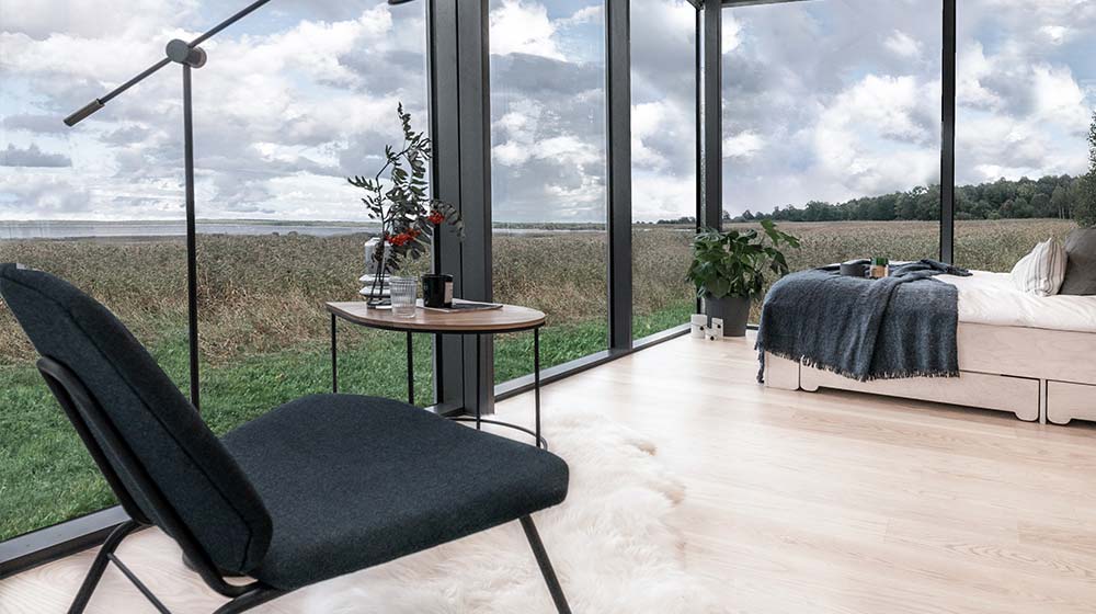Bright, minimalist interior of a mirror cabin with large glass windows overlooking nature, featuring a bed, chair, and small table with plants.