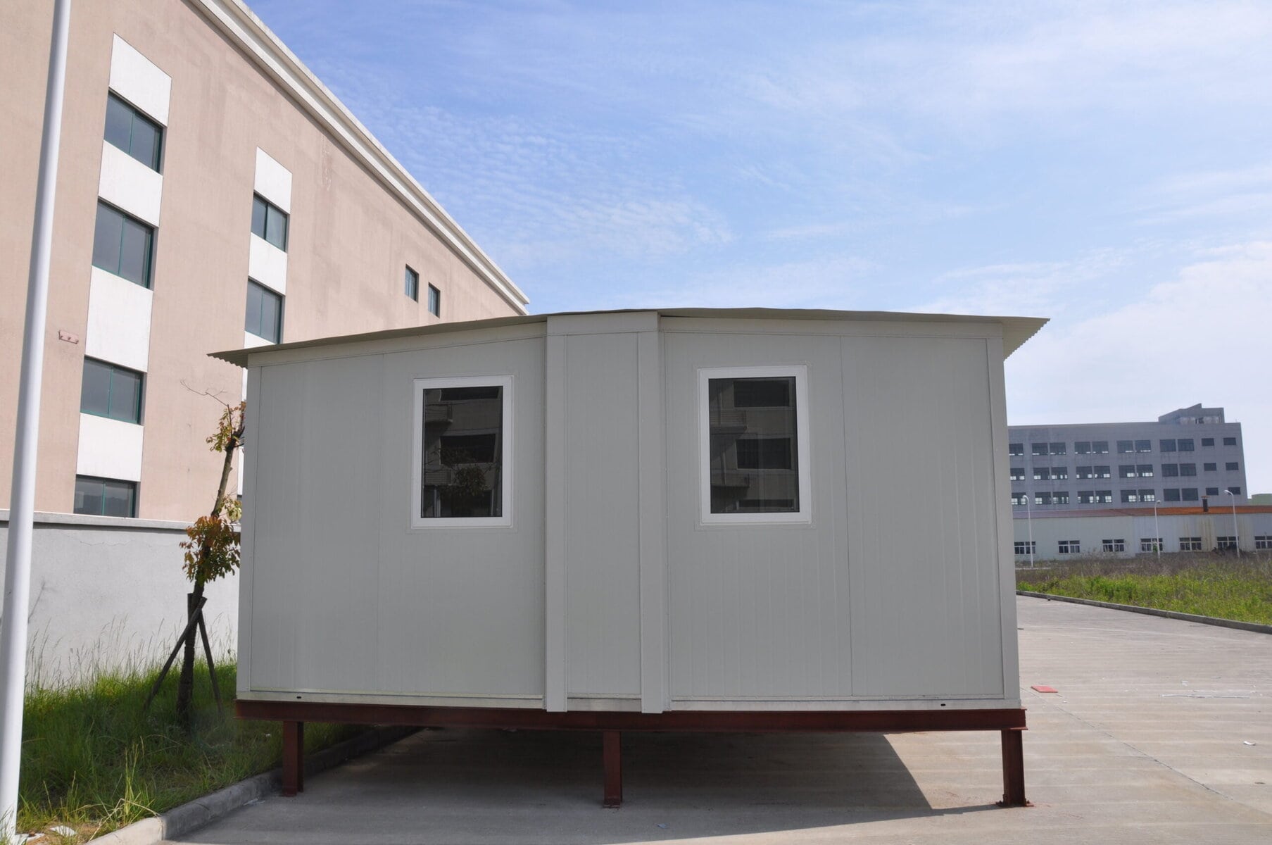 Modern tiny home on stilts with white exterior and two windows, set in an urban environment with buildings and a blue sky in the background.