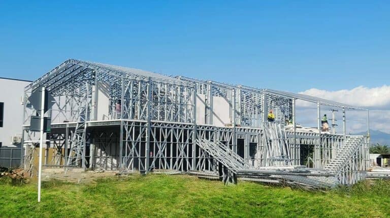 Steel frame building under construction on grass, workers on site, clear sky; prefab home techniques in Fiji shown.