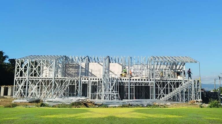 Workers build a steel-framed prefab home under a clear blue sky in Fiji, showing modern construction techniques.