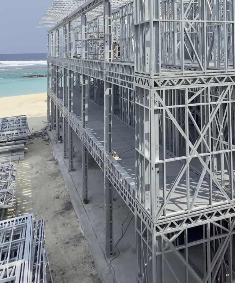 Steel frame building under construction by a sandy beach, turquoise water visible in the background.