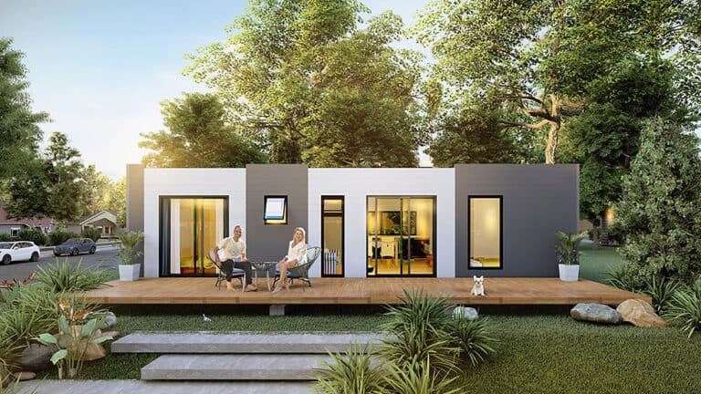 Two people sit on a deck by a modern house with big windows and trees around. A small dog sits close to them.