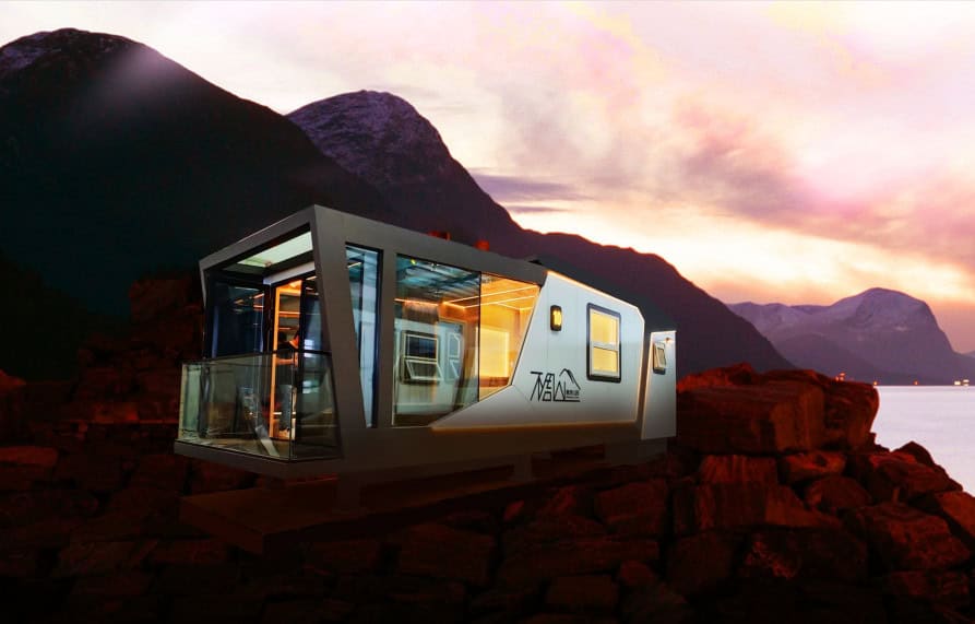A glass tiny house on rocky ground at dusk, with mountains and water in the background.