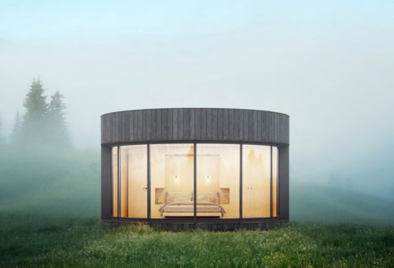 Modern circular cabin with glass walls, glowing interior, and a bed, set in a grassy, misty field with trees behind.