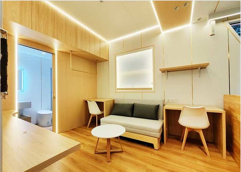 Minimalist room with wood floors, sofa, two desks, round table, and a bathroom seen through an open door.
