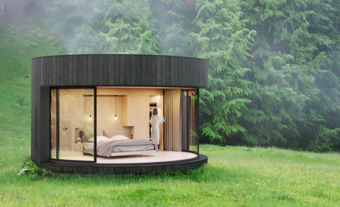 Circular cabin with tall windows on grass; person stands by bed inside, trees visible in the background.