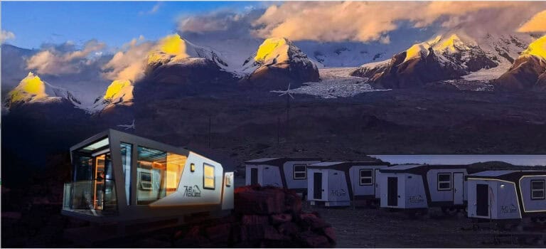 Modern modular cabins sit on rocky ground with snow-capped mountains and a glacier behind them under a partly cloudy sky.