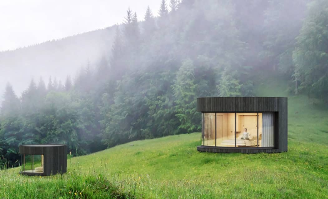 Two round black cabins with large windows sit on a grassy hill, surrounded by dense forest and mist.
