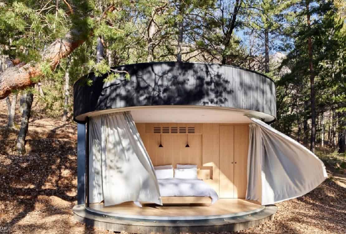 A round, modern cabin with open curtains and a visible bed sits among trees in a wooded area.