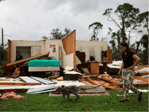 A woman walks a dog past a storm-damaged house, with debris scattered and parts of the structure still standing.