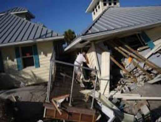 A person stands in debris between two houses with collapsed roofs and walls under a clear blue sky.