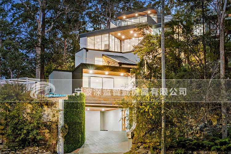 Modern multi-level house with large windows, outdoor lighting, trees, greenery, and a driveway to the garage.