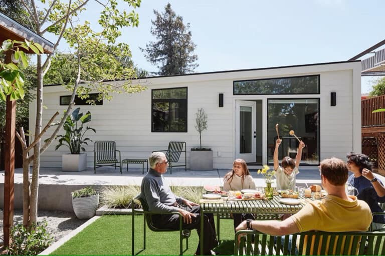 Five people eat and talk around an outdoor table in a backyard with a white guesthouse and lawn chairs in the background.