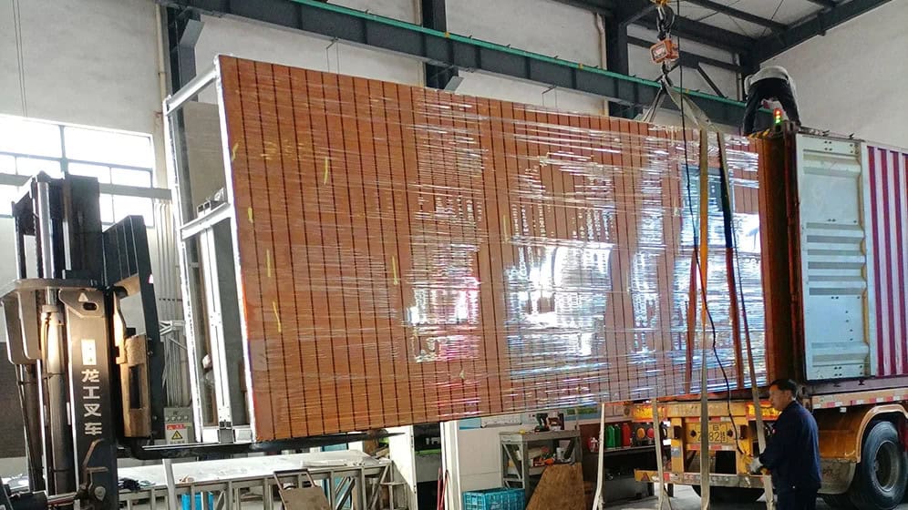 A forklift and crane load a large, wrapped object into a shipping container in a warehouse as two workers watch.