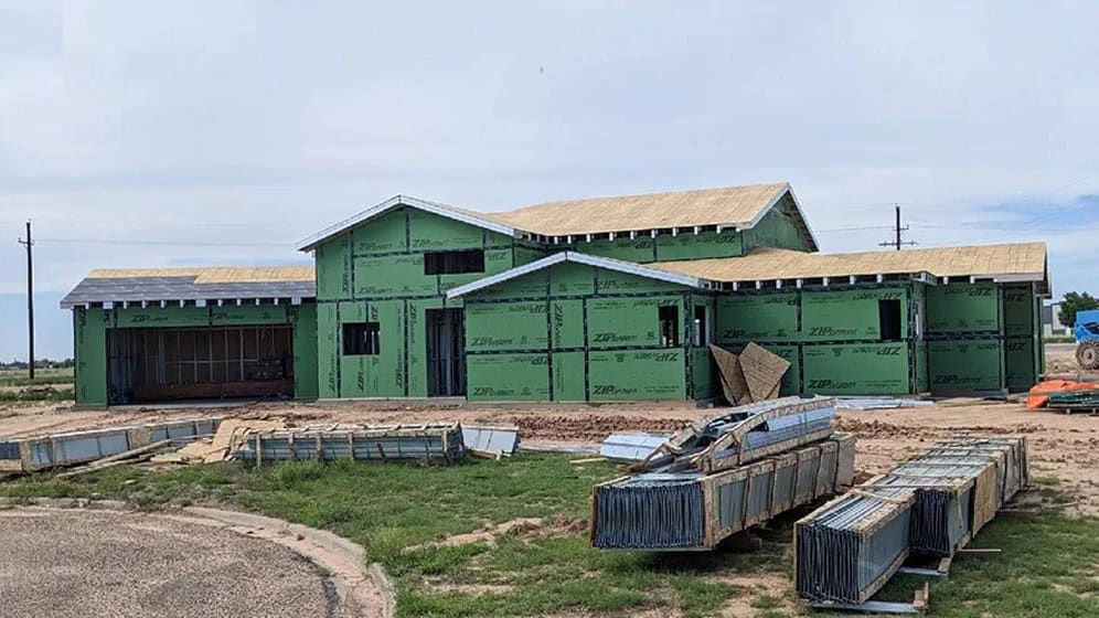 Two-story house under construction with green sheathing, building materials, and metal ducts scattered in front.