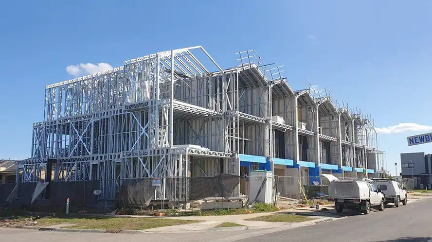 Steel-framed building under construction with scaffolding and nearby vehicles on a clear day.