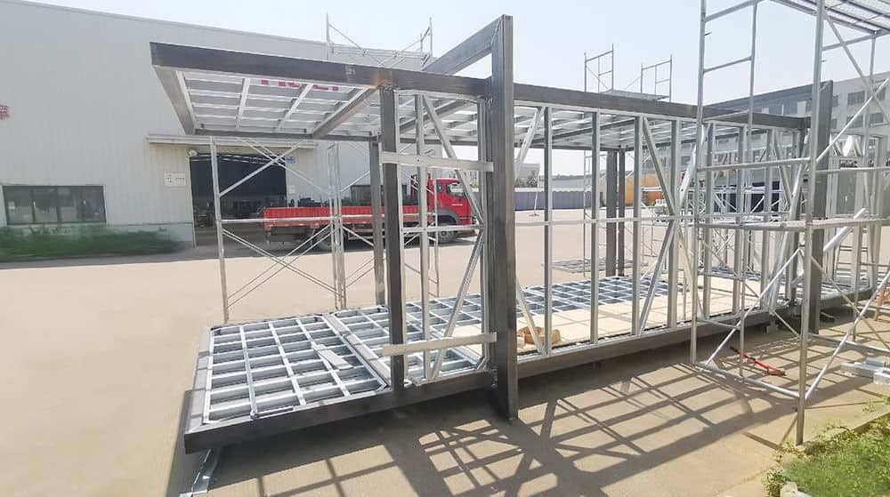 A steel modular building frame is being built outdoors, with scaffolding and a red truck in the background.