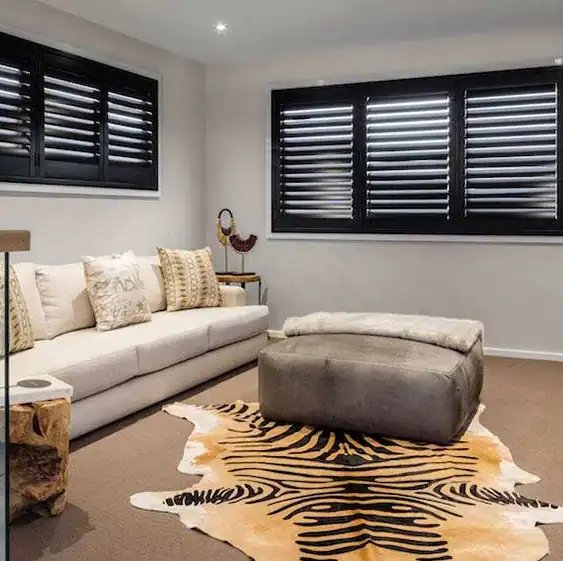 Modern living room with white sofa, patterned pillows, ottoman, animal print rug, side table, and black shuttered windows.