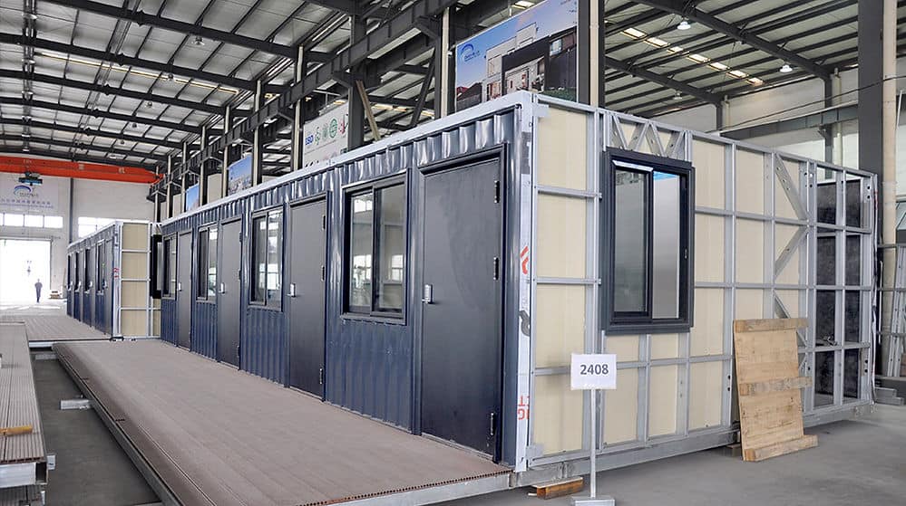A modular building with dark doors and windows is being built in a factory; steel framing is exposed on the right.