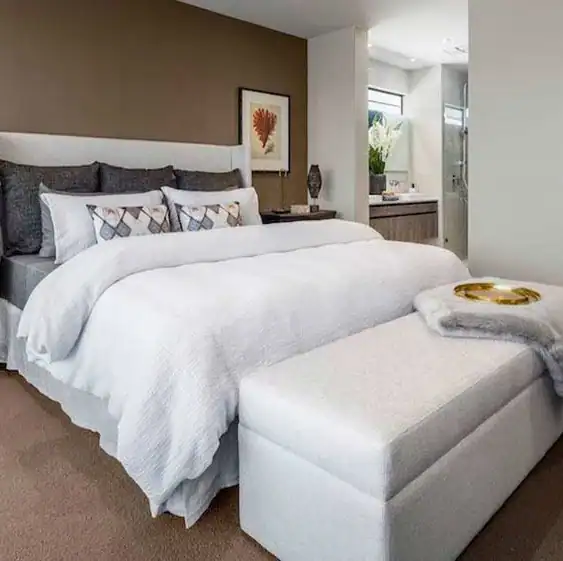 Modern neutral bedroom with a made bed, pillows, cushioned bench, and a view into the bathroom.