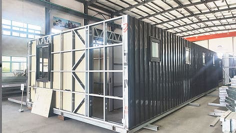 An indoor shipping container under construction with exposed framing, unfinished walls, and windows.