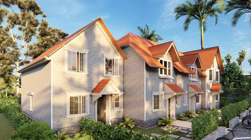 Modern townhouses with red roofs, white brick exteriors, and neat gardens among greenery and palm trees under a blue sky.