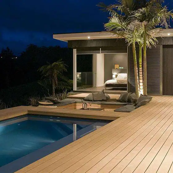 Modern deck with small pool, cushions, palm tree, and a lit bedroom behind large glass doors at night.