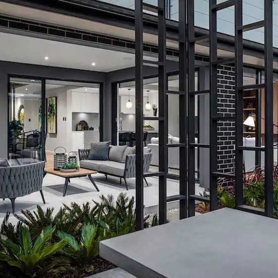 Modern patio with gray furniture and coffee table beside glass-walled living room, kitchen, and landscaped plants.