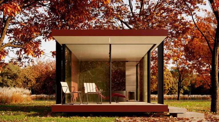Glass-walled cabin with patio chairs amid autumn trees and fallen leaves in a park-like setting.