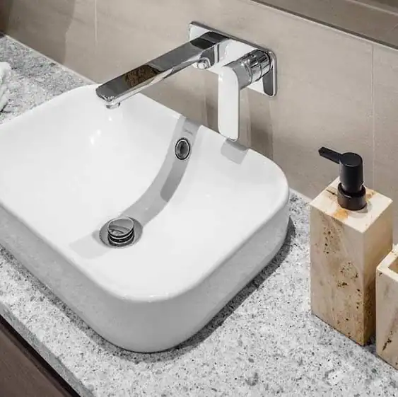 White rectangular sink with wall-mounted chrome faucet on gray countertop; soap dispenser at right.