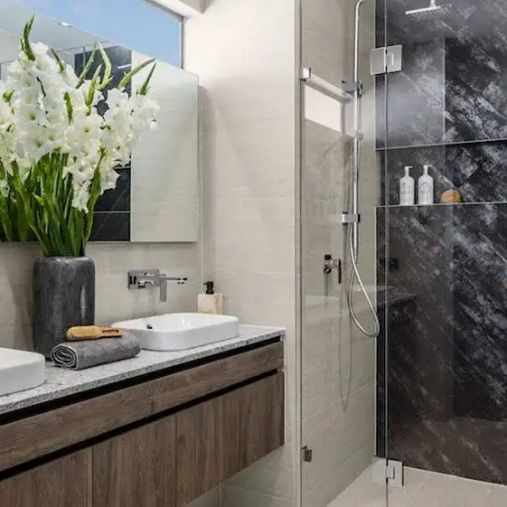 Modern bathroom with dual sinks, vase of white flowers, and glass shower with dark marble walls and shelves with bottles.