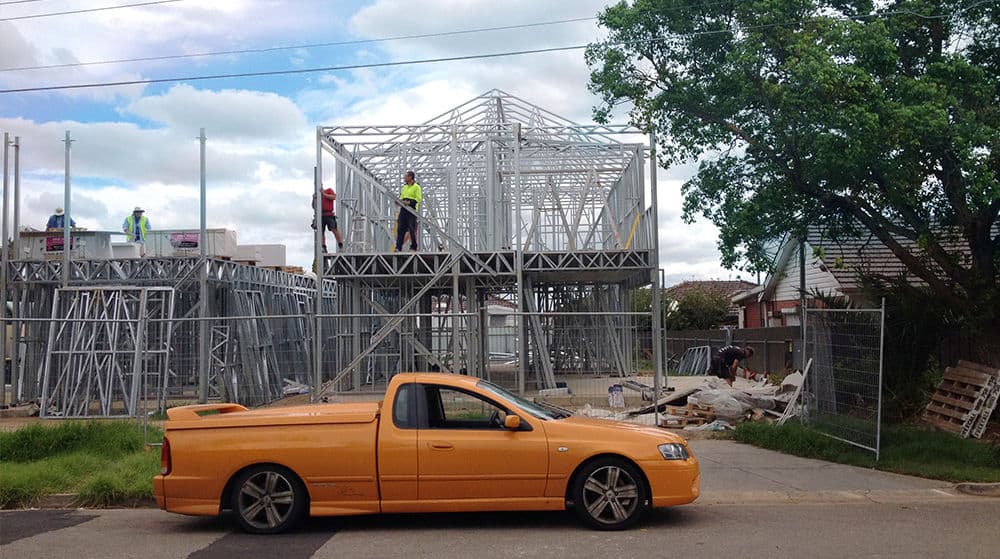 A yellow utility vehicle in front of a steel-framed house under construction, with workers and scattered materials nearby.