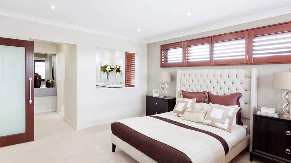 Modern bedroom with beige headboard, white and brown bedding, bedside lamps, wood shutters; bathroom seen through doorway.