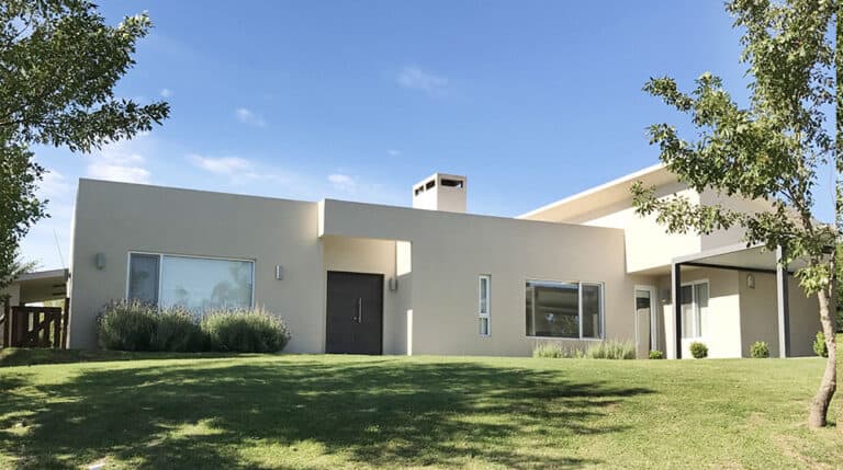 Single-story beige house with flat roof, large windows, and green lawn amid trees under a clear blue sky.