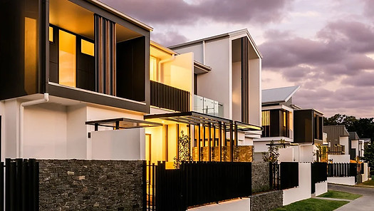 Modern two-story houses with large windows and warm lights, stone and metal accents, beneath a cloudy evening sky.