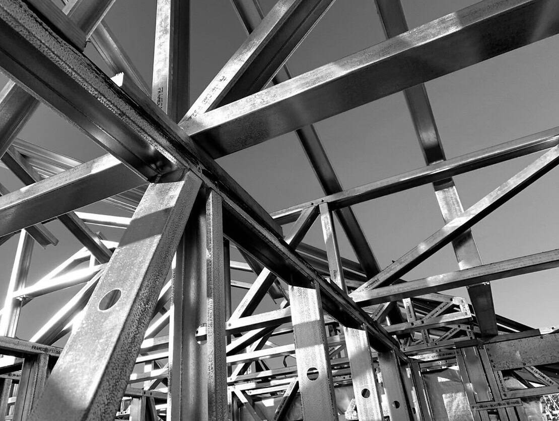 Black and white photo of intersecting steel beams forming a building framework under construction against a clear sky.