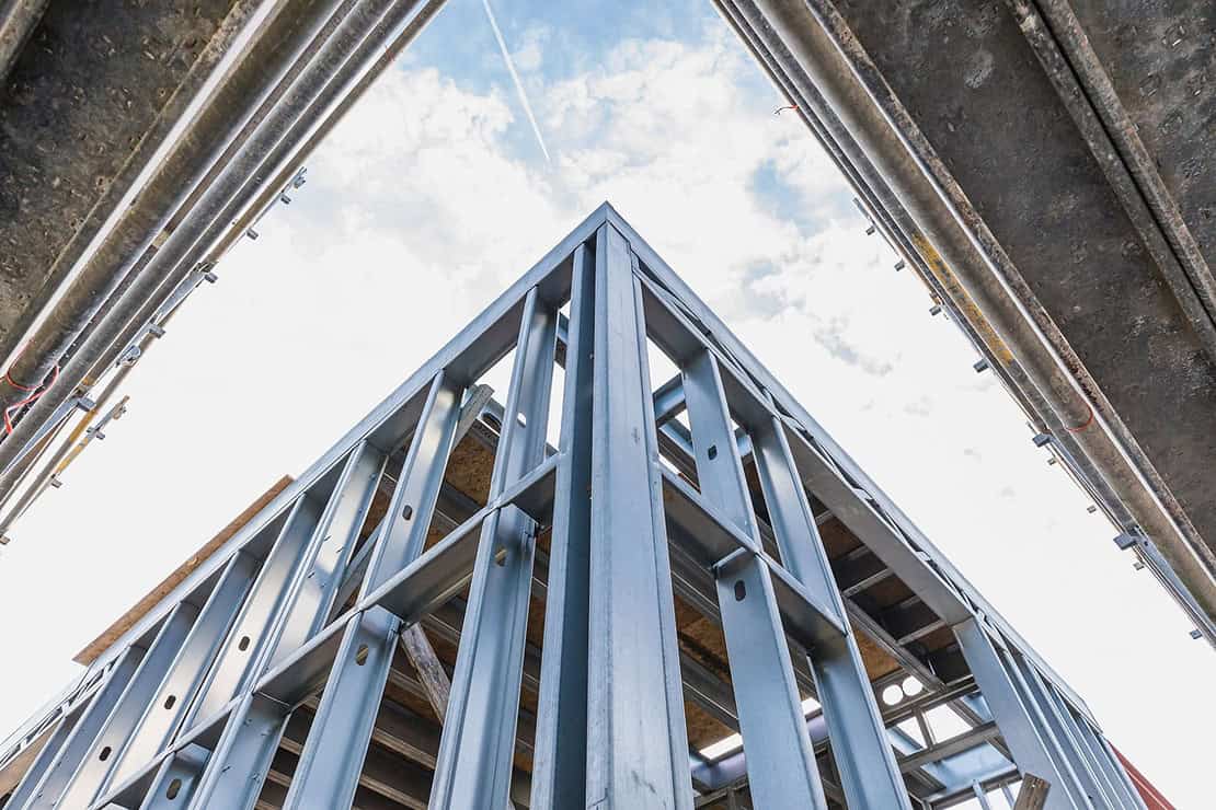 Steel frame of a building under construction seen from below, with blue sky and clouds in the background.