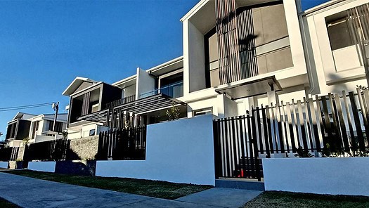 Modern two-story townhouses with large windows, balconies, and black metal fences line a sidewalk under a blue sky.