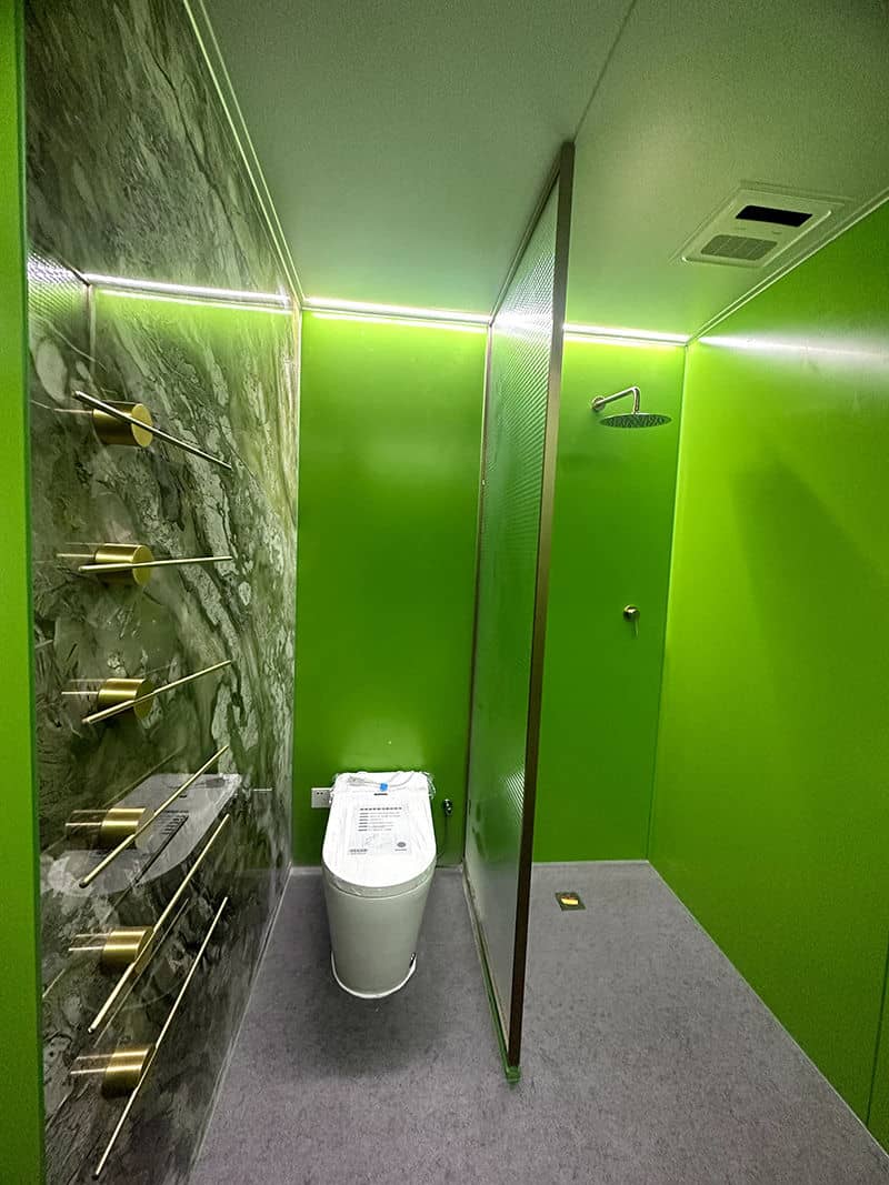 Modern bathroom with bright green walls, toilet, rainfall shower, marble accent wall, and gold towel bars.