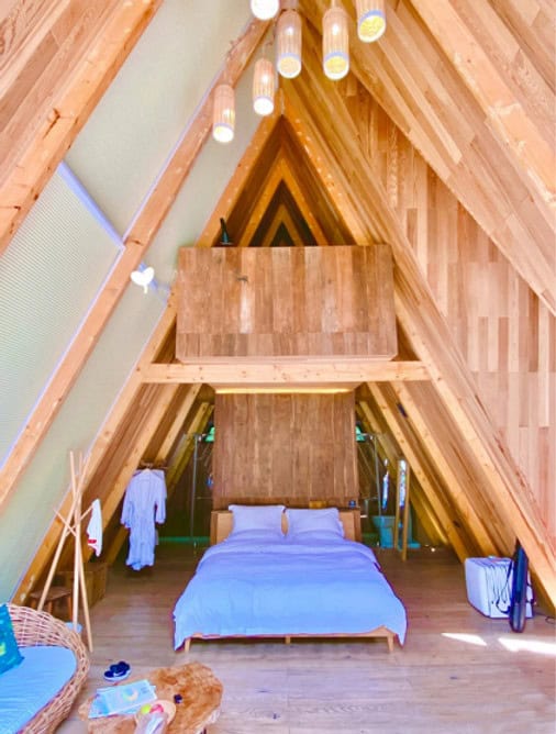 A-frame cabin bedroom with double bed, loft, hanging lights, and minimalist decor.