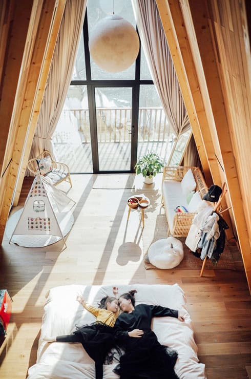 A woman and child lie on a bed in a sunlit A-frame room with large windows, cozy decor, and snowy views outside.