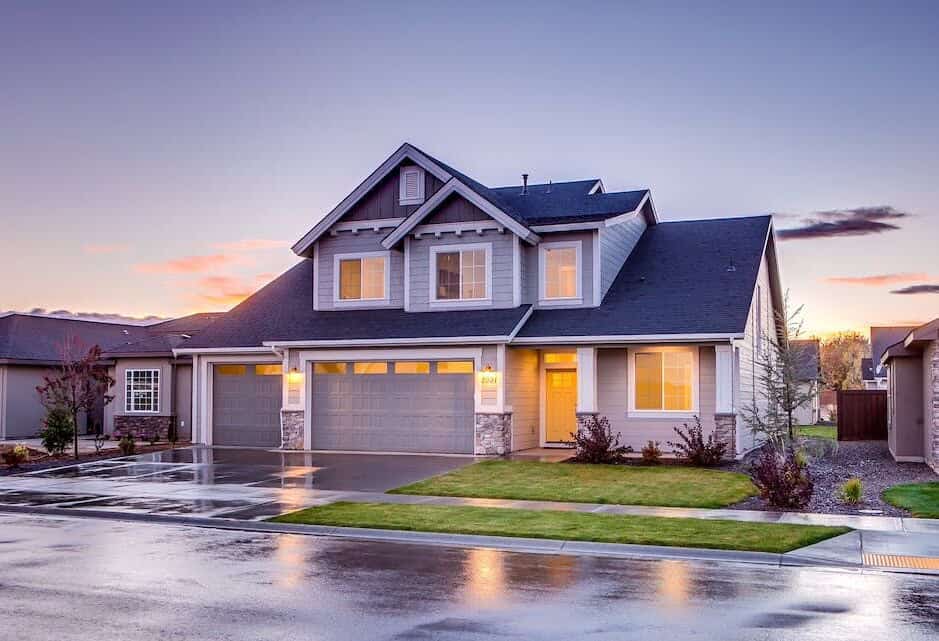 Modern two-story house with three garage doors, glowing windows, and a wet driveway at sunset.