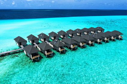 Overwater bungalows with dark roofs stand above turquoise water, linked by wooden walkways.