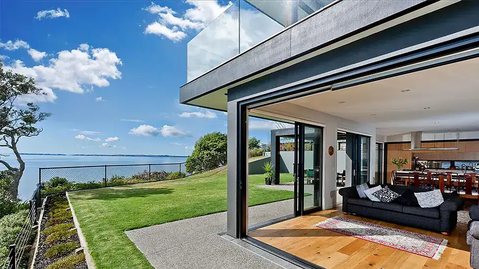 Modern house with glass doors to a grassy yard, overlooking water and blue sky. Contemporary interior visible.